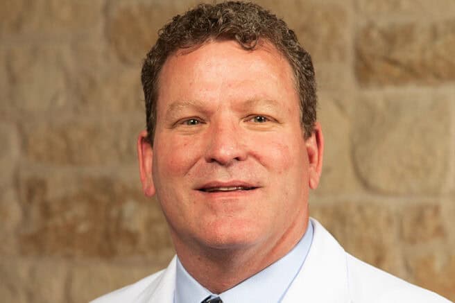 A healthcare professional in a white coat smiling against a brick wall background.