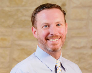 A healthcare professional smiling in a light blue shirt and tie against a beige brick wall backgroun.