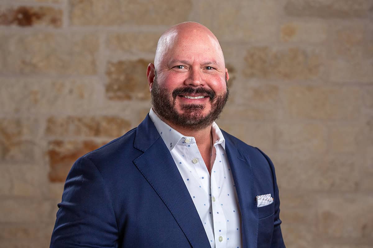 A healthcare professional in a navy suit smiling against a brick wall background.