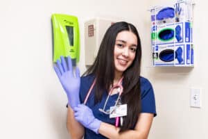 Nurse in scrubs and gloves smiling in a healthcare setting.
