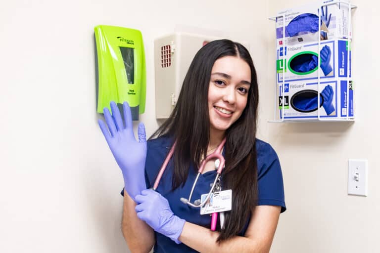Nurse in scrubs and gloves smiling in a healthcare setting.