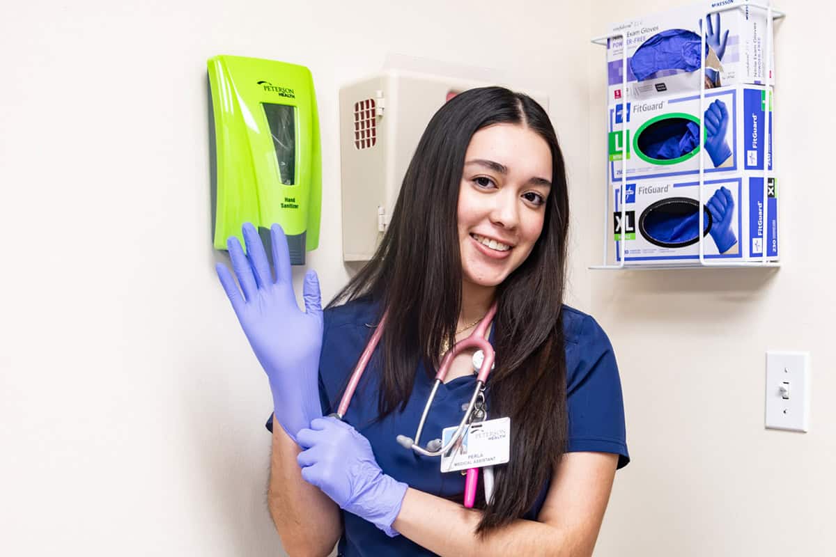 Nurse in scrubs and gloves smiling in a healthcare setting.