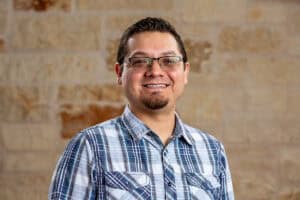 Smiling man with glasses and plaid shirt in front of brick wall.
