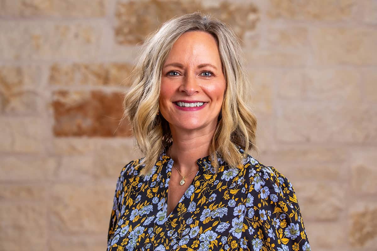 Smiling woman with blonde hair in front of a brick wall, wearing a floral blouse.