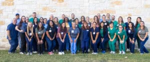 Group of healthcare professionals outdoors in front of a stone wall, smiling and wearing scrubs.