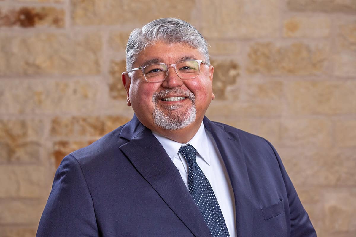 Smiling middle-aged man in a navy suit and tie against a brick wall background.